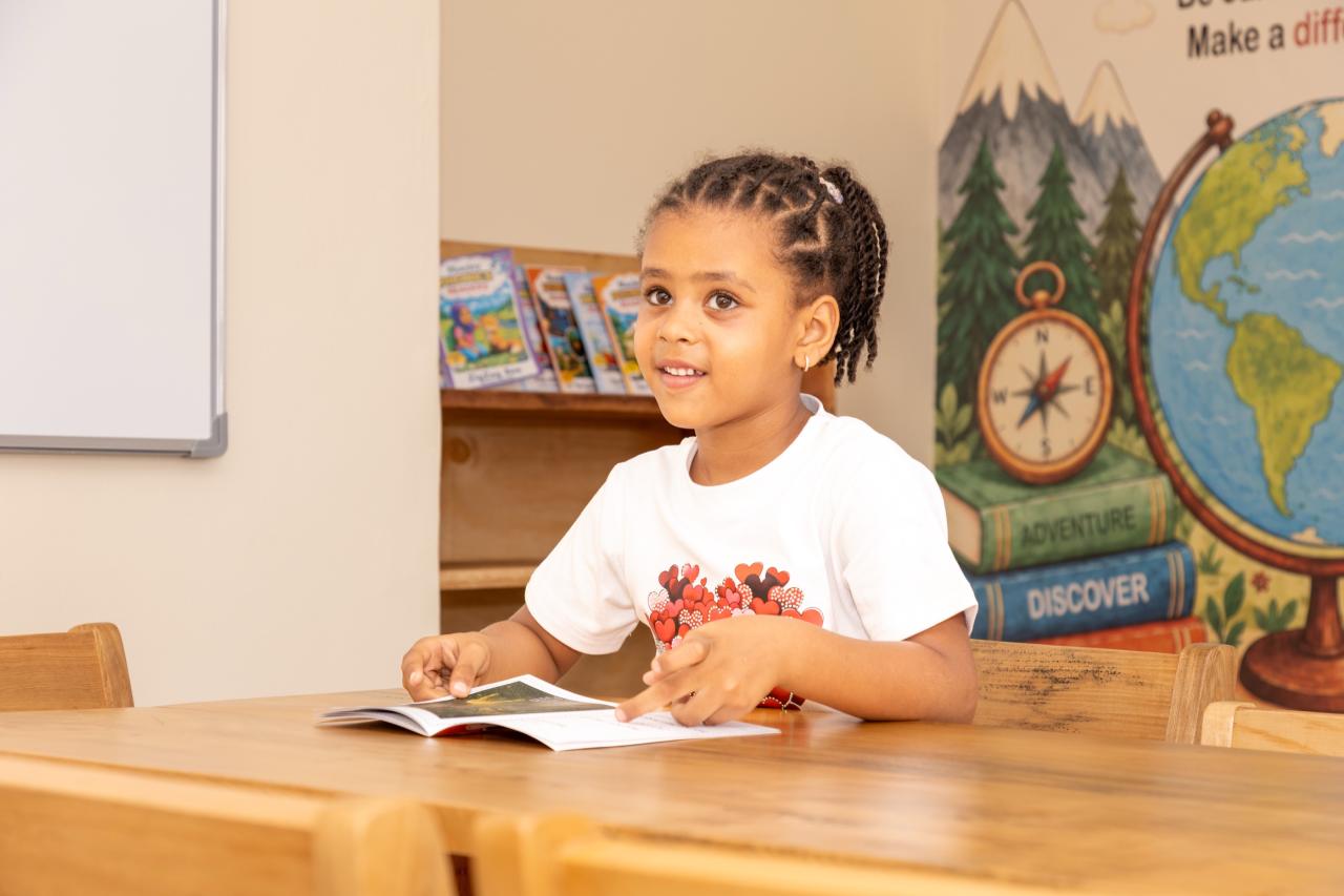 A smiling girl reading at a classroom table in front of a globe and an explorer mural