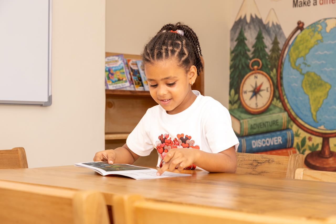 A smiling girl reading at her classroom table beside a globe and an adventure mural